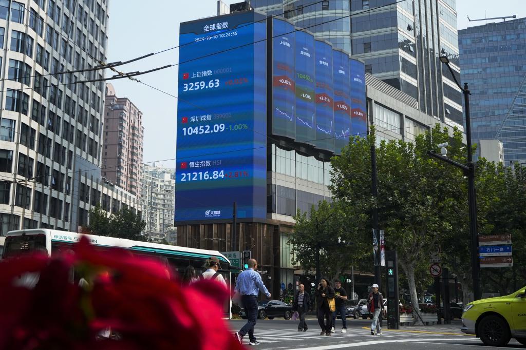 Pedestrians pass by an electronic board displaying Shanghai, top, Shenzhen, center, and Hang Seng, bottom, shares trading indexes at a commercial office building in Shanghai