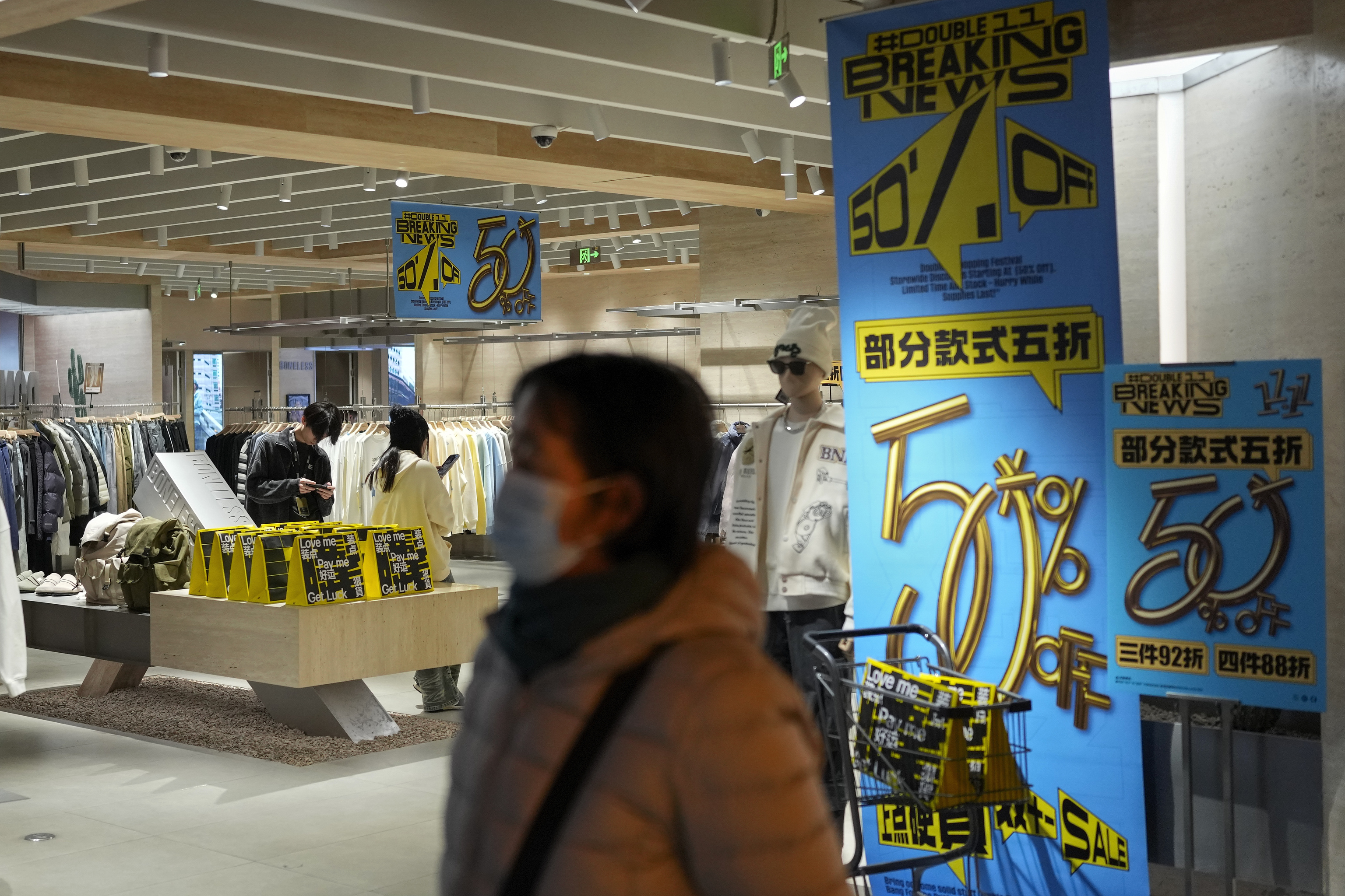 A masked woman walks at a fashion boutique displaying posters to promote Singles' Day discounts at a shopping mall.