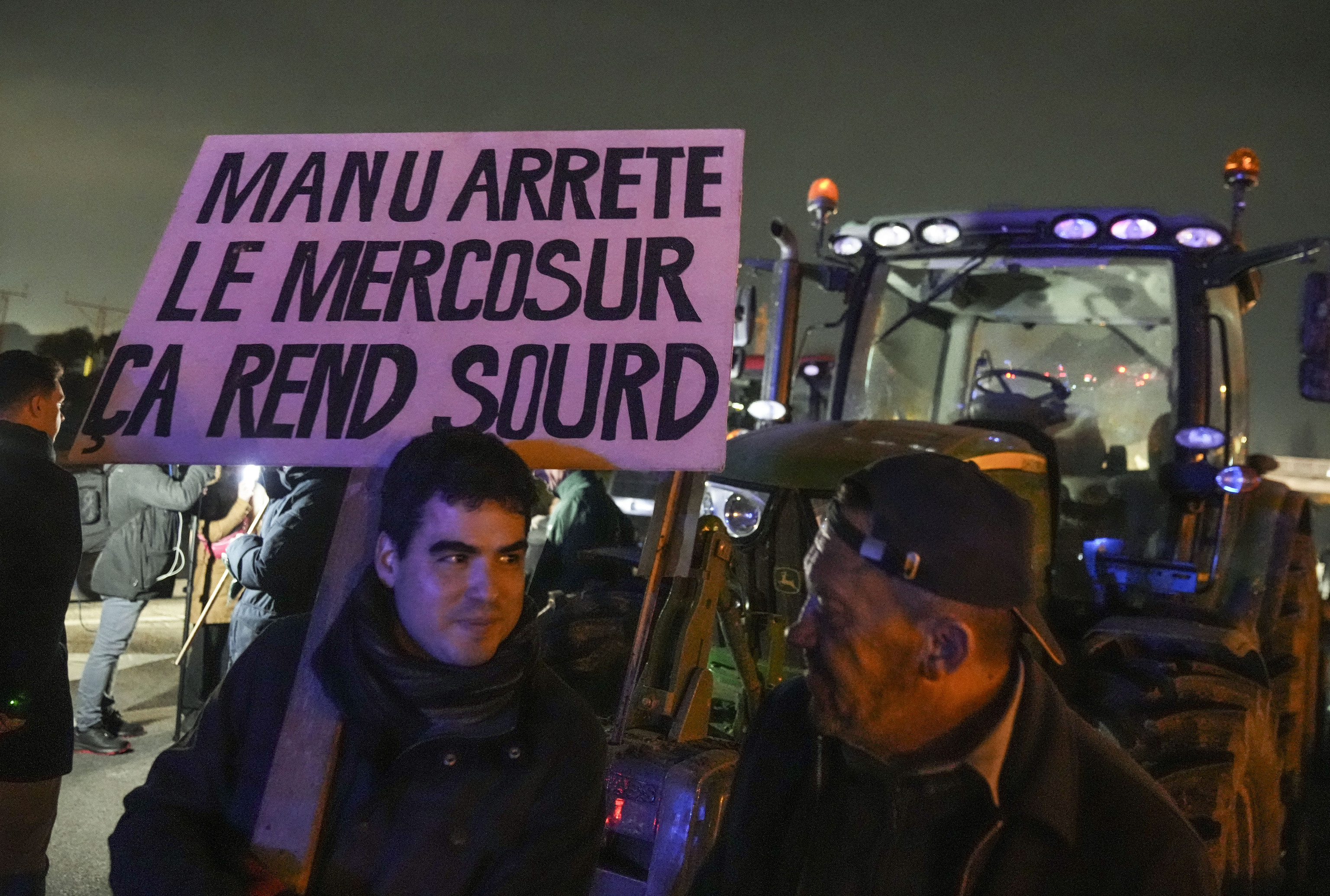 A farmer holds a placard that reads, "Macron stop the Mercosur it makes you deaf " as he stands next to tractors on a blocked highway.