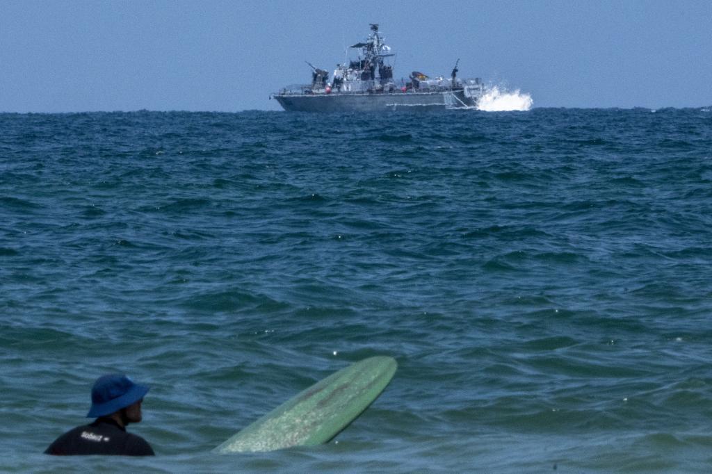 A surfer waits for wave while an Israeli military naval ship patrols the Mediterranean sea off the coast of Hadera