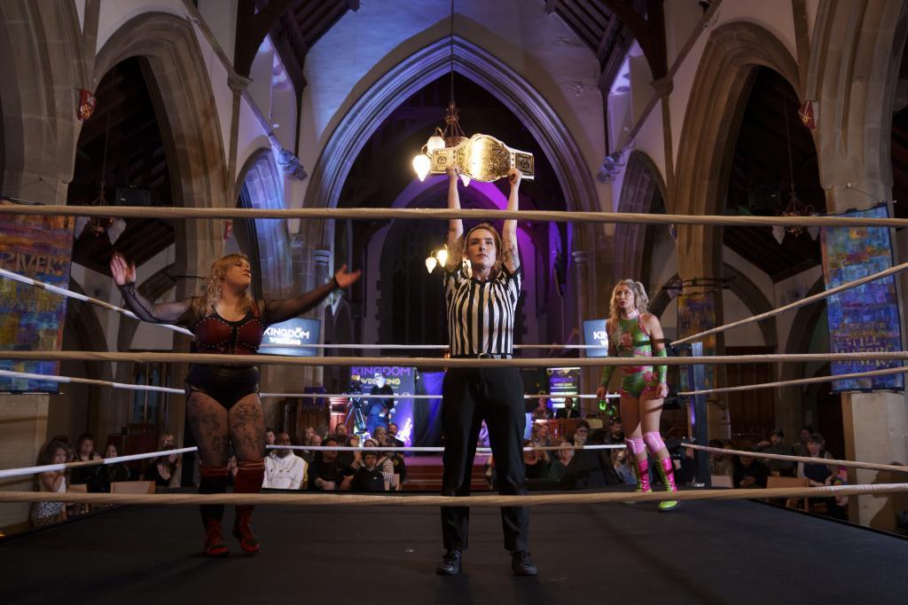 Referee Katie Crosby holds up the contested belt before Kiara's bout against Scarlett, left, during a Kingdom Wrestling show at St Peter's Church in Shipley