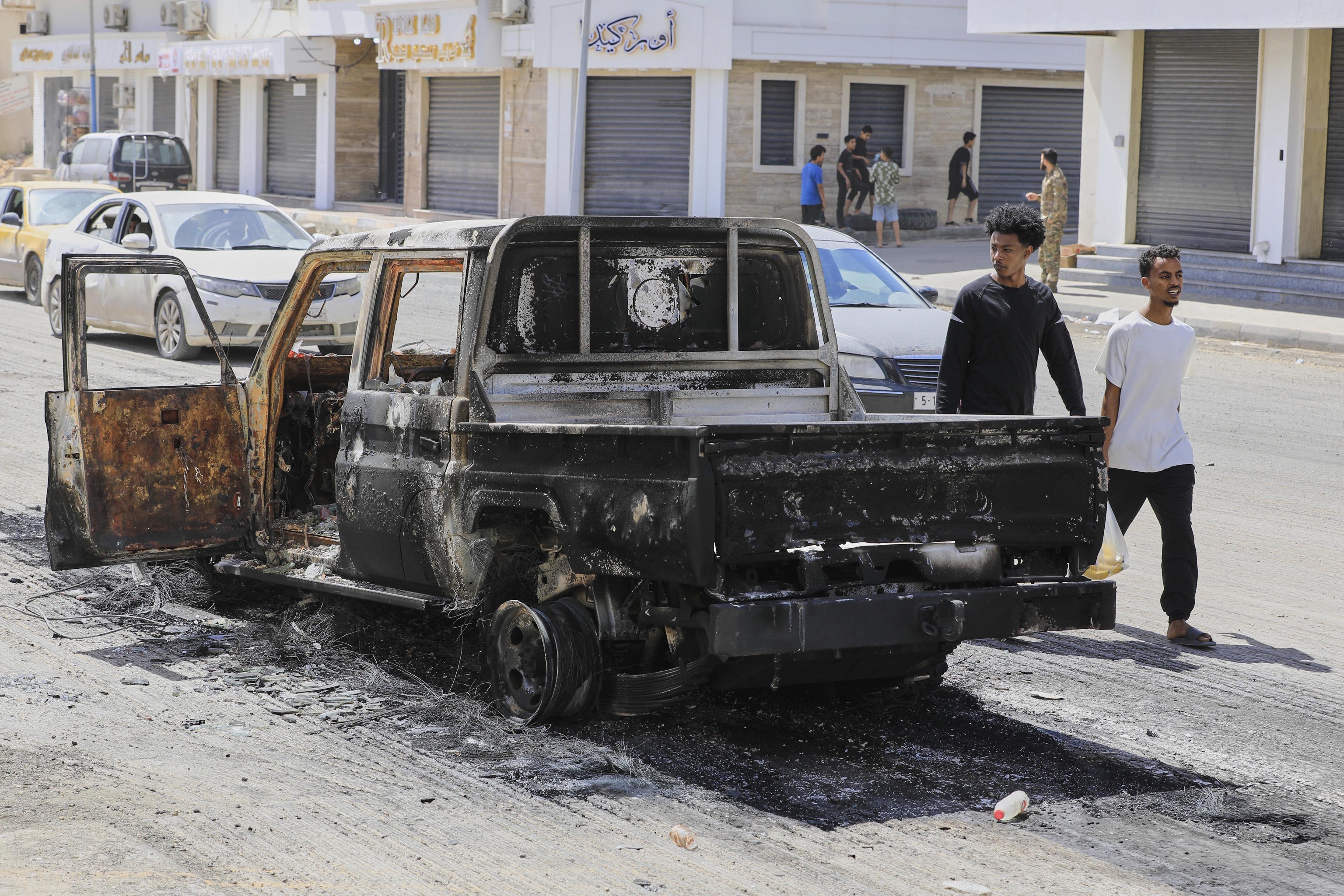 Libyans walk past a burnt vehicle after Monday evening clashes between armed militias in Tripoli.