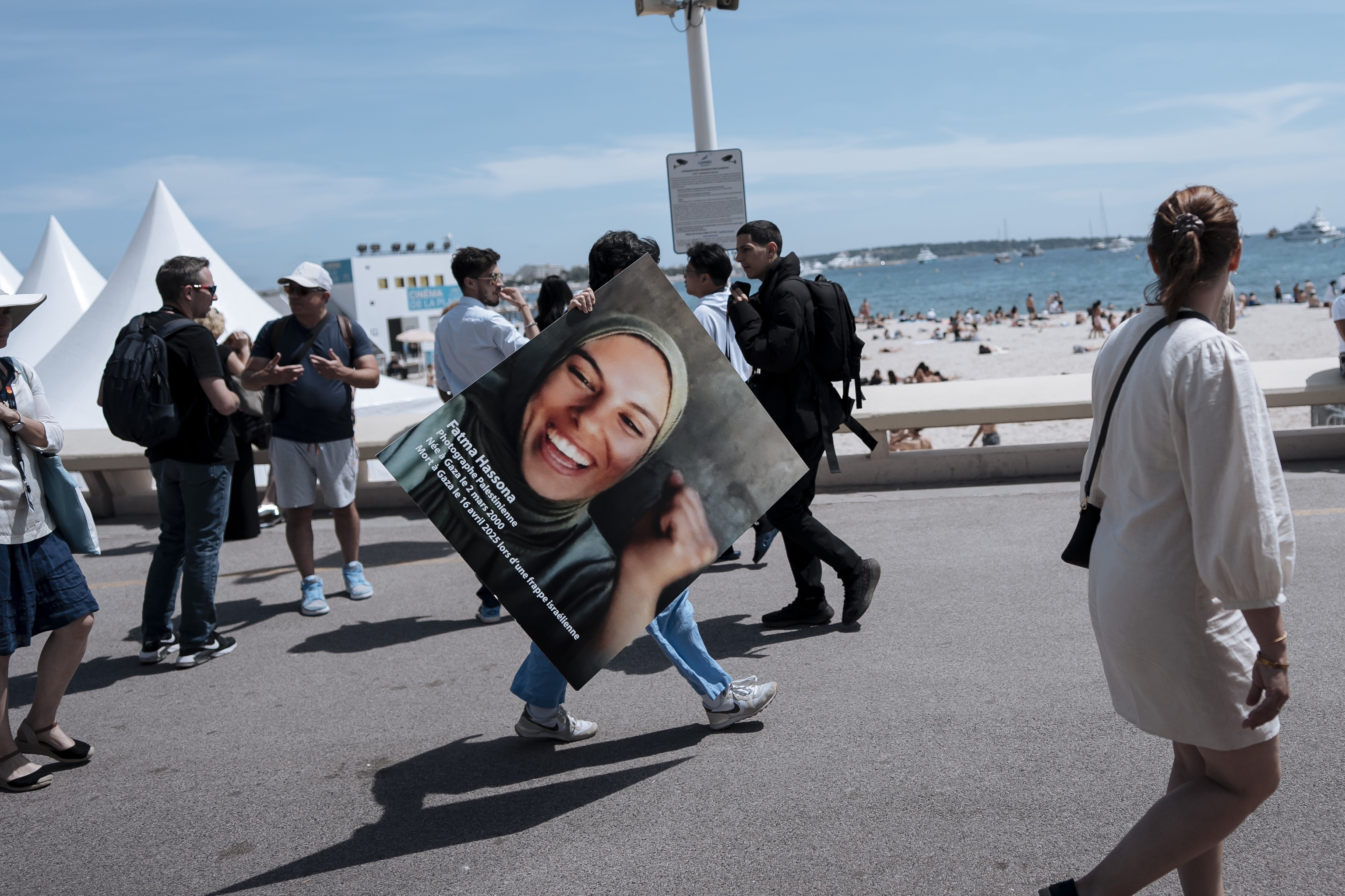 A person walks along the Boulevard de la Croisette holding a poster which reads 'Fatma Hassona Palestinian Photographer born in Gaza on 2nd March 2000 died in Gaza on April 16th 2025 during an Israeli strike.