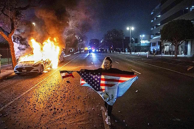 A woman walks next to a burning car during immigration raid protests in Los Angeles.