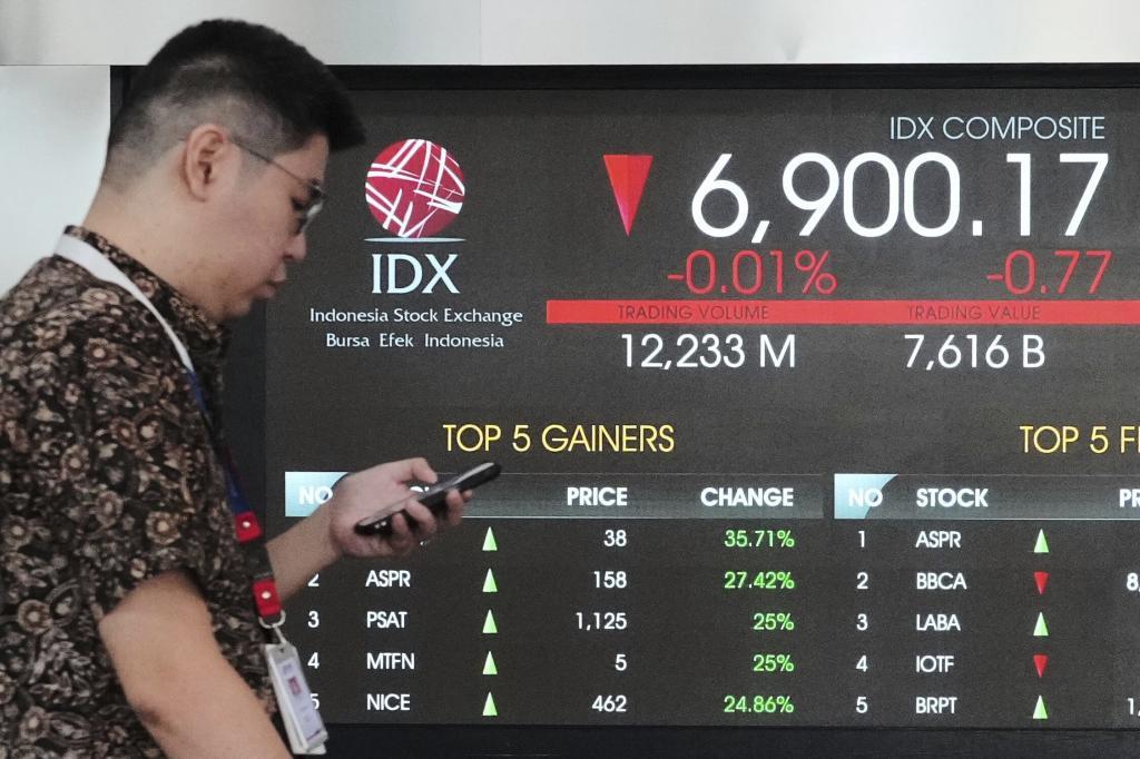 A man checks his mobile phone as he walks past an electronic board displaying stock prices and Jakarta Stock Exchange Composite Index
