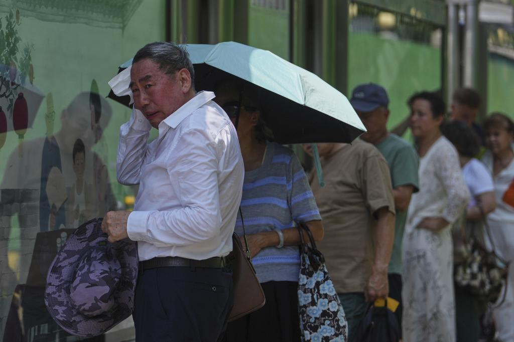 A man wipes sweat as commuters stand in shade at a bus stand waiting for their buses