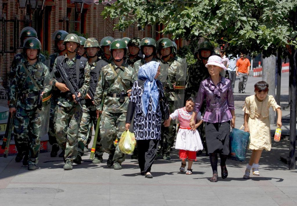 Uighur woman and children walk near a platoon of paramilitary police