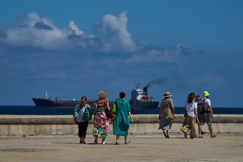 Tourists walk along the seafront promenade while a tanker exits Havana