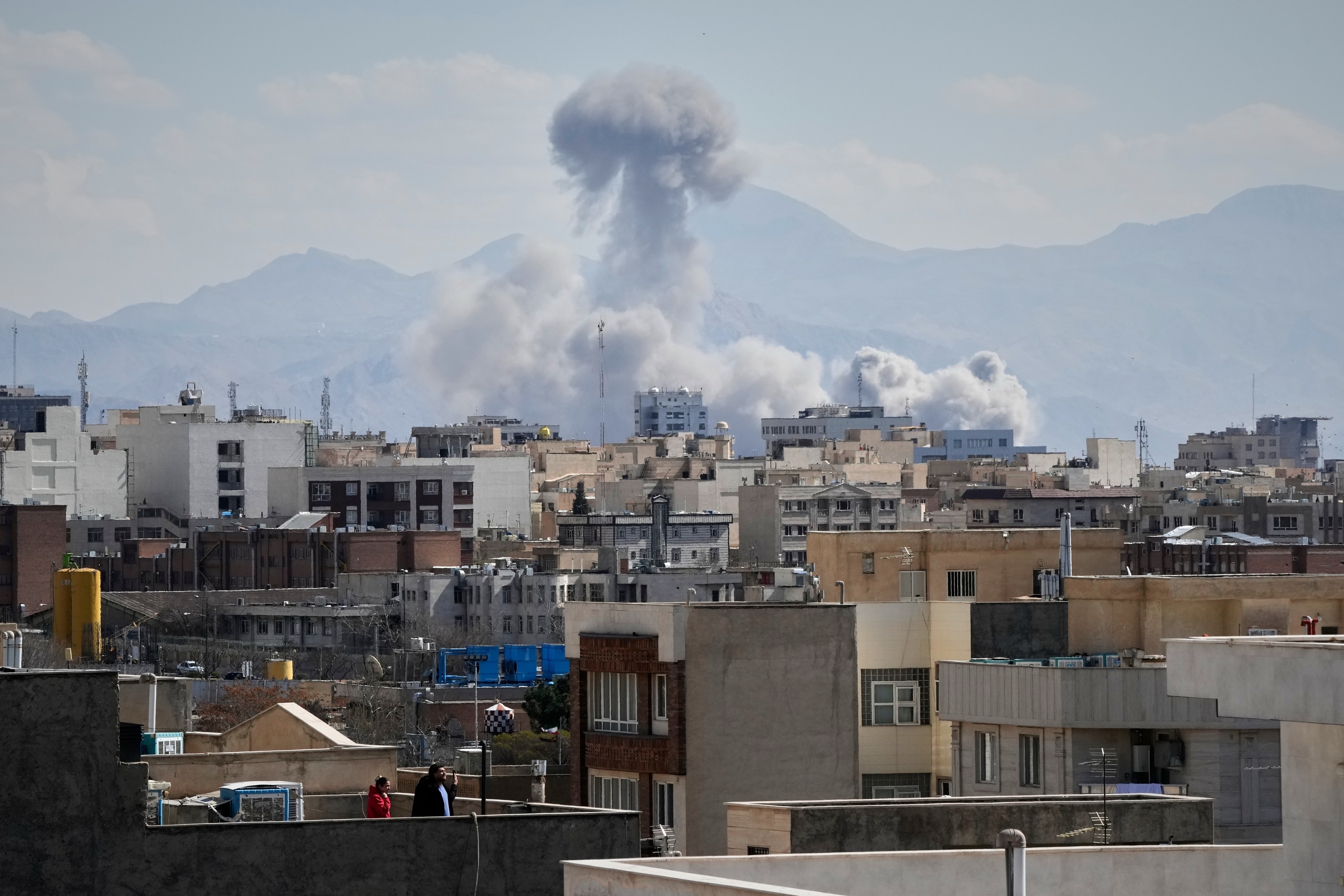People watches from a rooftop as a plume of smoke rises after a strike in Tehran, Iran.