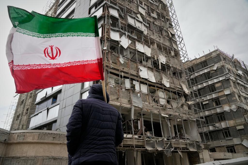 A man holds an Iranian flag as he looks at the damaged fa�ade of Gandhi Hospital