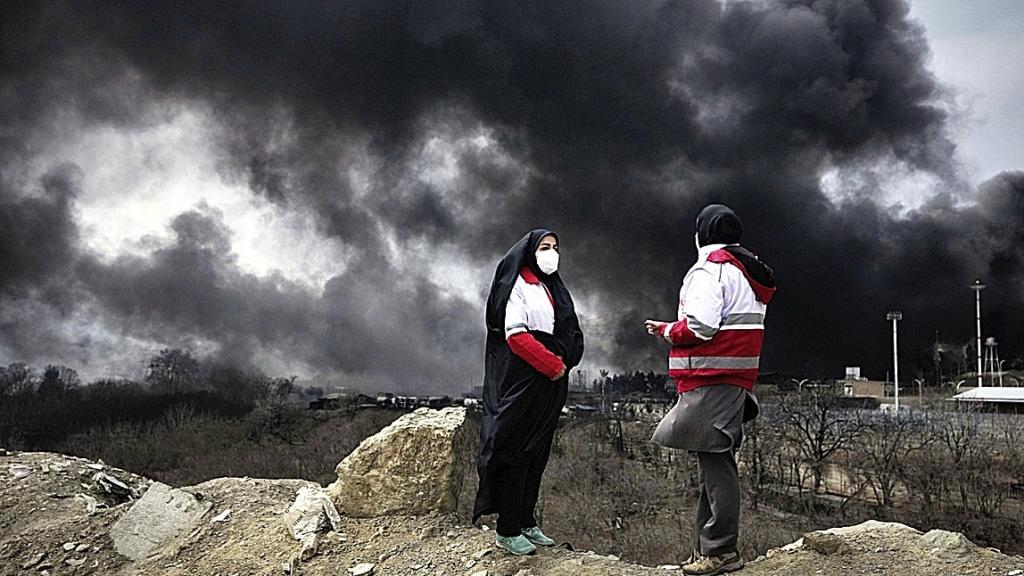 Two women from the Iranian Red Crescent Society stand as a thick plume of smoke from a U.S.-Israeli strike on an oil storage facility
