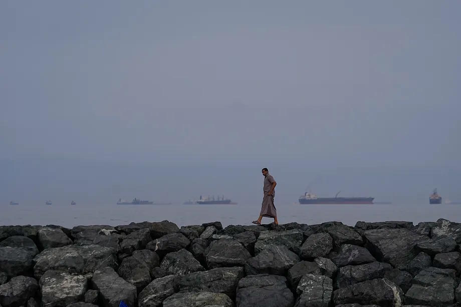 A man walks along the seashore as oil tankers and cargo ships line up in the Strait of Hormuz.