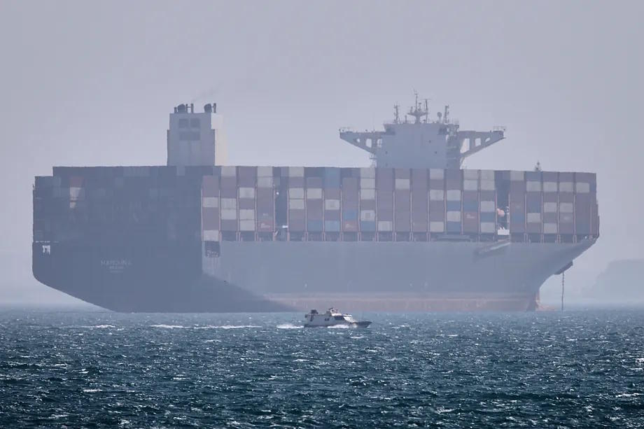 A container ship remains anchored while a small motorboat passes in the foreground in the Strait of Hormuz.