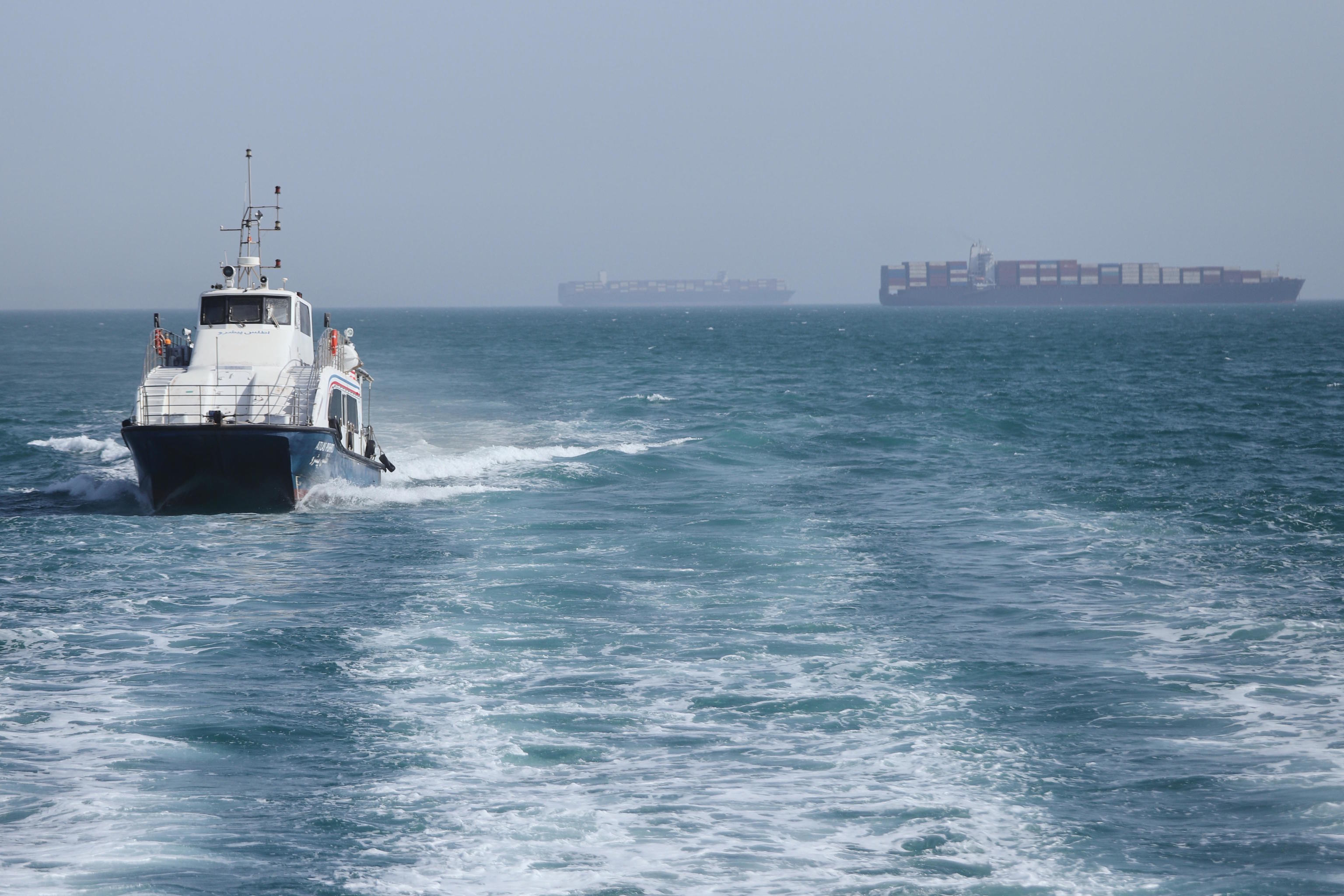 A patrol boat sails while cargo ships remain anchored in the Strait of Hormuz.