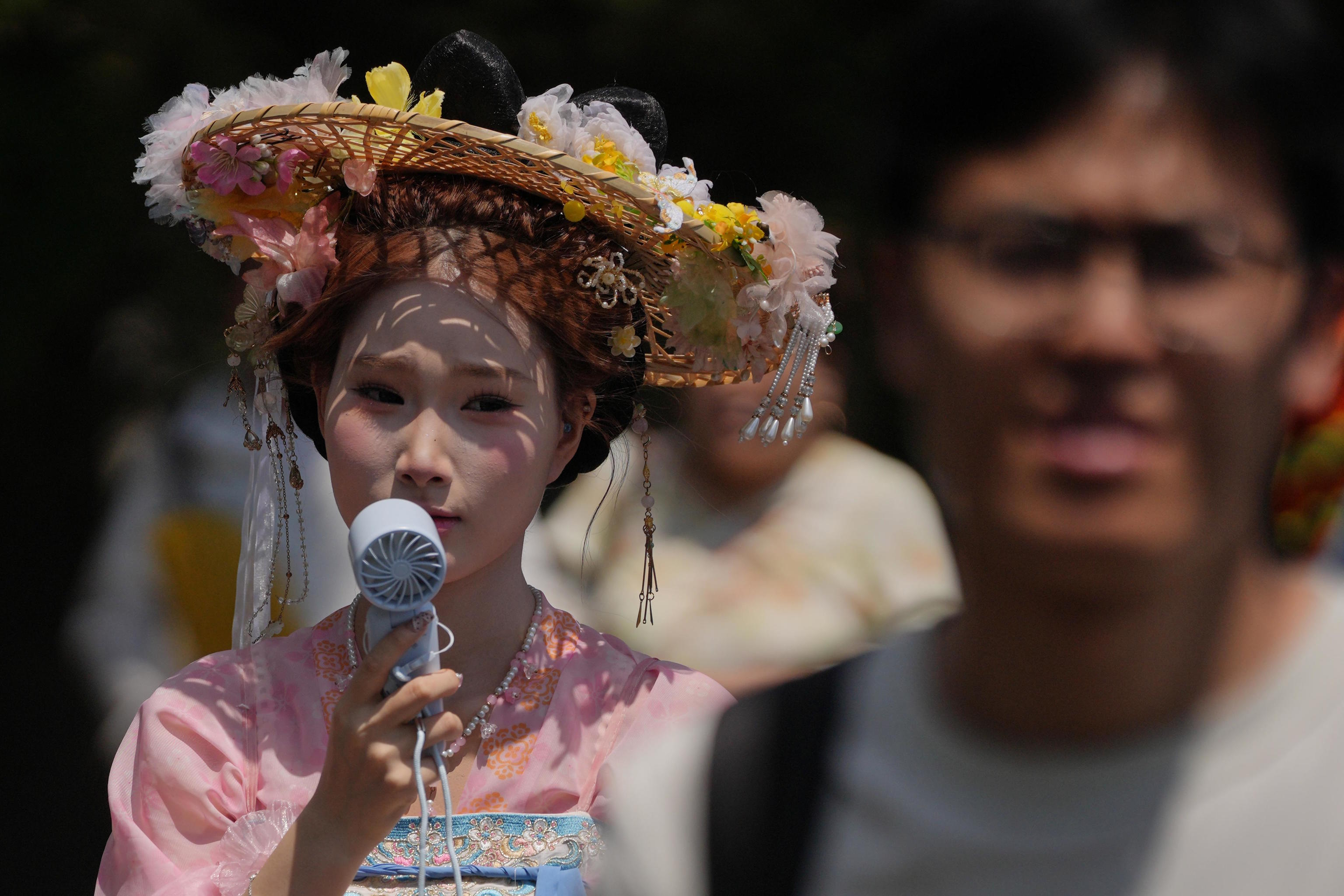 A woman wearing a hat with flowers and traditional costume cools herself with a portable electric on a hot day, in Beijing.